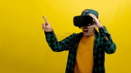 Un joven asiático con auriculares VR señala con el dedo hacia adelante, interactuando con una simulación de realidad virtual. Aislado sobre un fondo amarillo