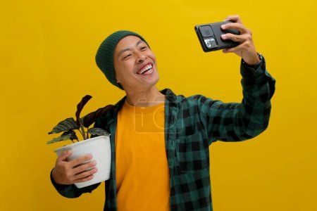 Ein fröhlicher asiatischer Mann in Mütze und lässiger Kleidung macht ein Selfie mit seiner gesunden Zimmerpflanze Calathea (Calathea ornata) in einem weißen Topf. Vereinzelt auf gelbem Hintergrund.