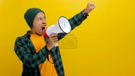 Téléchargez les photos : Un militant asiatique coiffé d'un bonnet crie et crie dans un mégaphone, exigeant des changements et protestant pour les droits de l'homme ou la justice en mettant l'accent sur la liberté d'expression. Isolé sur fond jaune - en image libre de droit