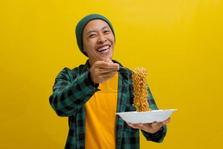 Hombre asiático en un gorro y ropa casual disfruta sorbiendo fideos instantáneos usando tenedor. Aislado sobre un fondo amarillo.