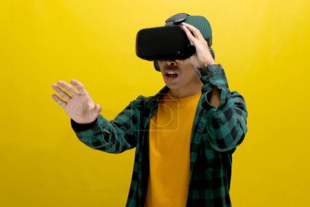 Emocionado hombre asiático con un casco VR extiende la mano, aparentemente interactuando con un objeto virtual en un juego de realidad virtual. Aislado sobre un fondo amarillo