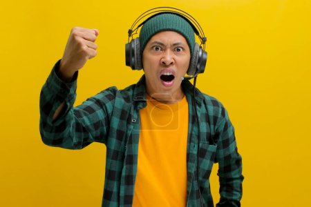 Furioso hombre asiático en un gorro y ropa casual, aprieta el puño mientras escucha música o podcast en los auriculares. Aislado sobre un fondo amarillo.