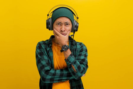 Hombre asiático en un gorro y ropa casual, profundo en el pensamiento con expresión pensativa mientras escucha música en los auriculares. Aislado sobre un fondo amarillo.