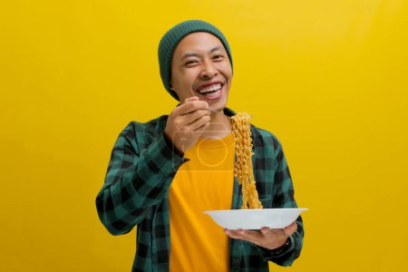 Homme asiatique dans un bonnet et des vêtements décontractés aime grignoter nouilles instantanées à l'aide de fourchette. Isolé sur fond jaune.