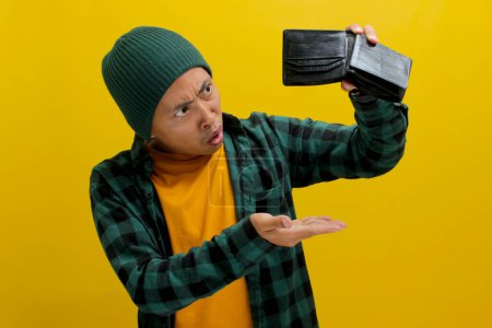 Dismayed Asian man stares at his empty wallet. Isolated on a yellow background. Financial hardship, budgeting, impulse spending, and saving concept.