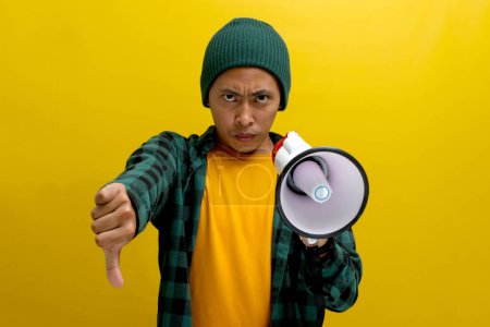 Displeased Asian man in a beanie hat holds a megaphone with an angry face, showing a thumbs-down to express disapproval or dissatisfaction, conveying rejection. Isolated on a yellow background