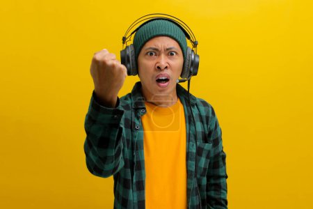 Homme asiatique furieux dans un bonnet et des vêtements décontractés, serre son poing tout en écoutant de la musique ou podcast sur écouteurs. Isolé sur fond jaune.