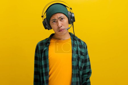 Hombre asiático en un gorro y ropa casual, profundo en el pensamiento con expresión pensativa mientras escucha música en los auriculares. Aislado sobre un fondo amarillo.