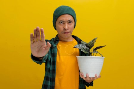 Asian man holds up a healthy Pin-stripe Calathea (Calathea ornata) houseplant in a white pot and makes a stopping gesture with his other hand, as if advising against bad houseplant care tips
