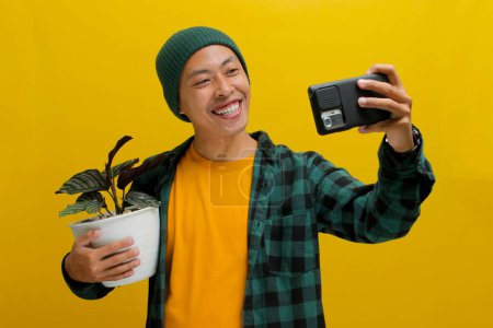 Ein fröhlicher asiatischer Mann in Mütze und lässiger Kleidung macht ein Selfie mit seiner gesunden Zimmerpflanze Calathea (Calathea ornata) in einem weißen Topf. Vereinzelt auf gelbem Hintergrund.