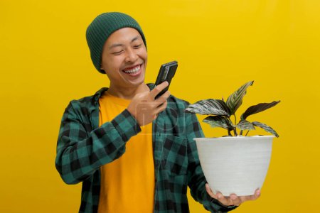 Cheerful Asian man in a beanie and casual clothes snaps a photo of his healthy Pin-stripe Calathea (Calathea ornata) houseplant in a white pot using his phone. Isolated on a yellow background.