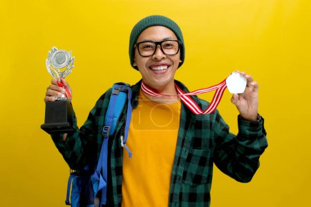 Happy young Asian student with a backpack, wearing eyeglasses, a beanie hat, and a casual shirt, shows off his medal and silver champion trophy, celebrating success. Isolated on a yellow background