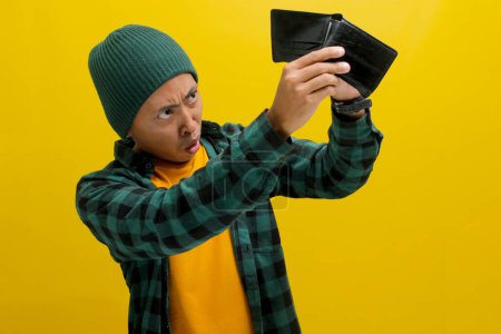 Un asiático consternado mira fijamente su cartera vacía. Aislado sobre un fondo amarillo. Dificultad financiera, presupuesto, gasto impulsivo y concepto de ahorro.