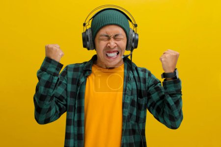 Furious Asian man in a beanie and casual clothes, clenches his fist while listening to music or podcast on headphones. Isolated on a yellow background.