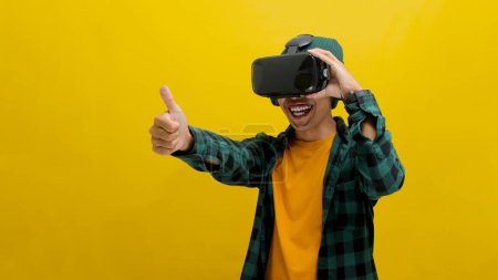 Happy Asian man wearing a VR headset gives a thumbs up, clearly enjoying his virtual reality experience. Recommends VR concept. Isolated on a yellow background