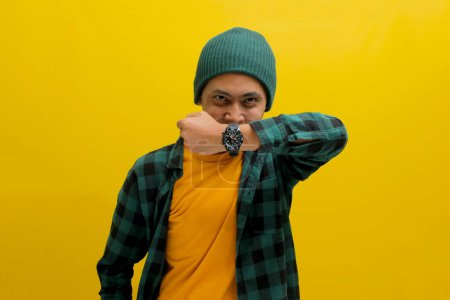 Impatient Asian man in a beanie and casual shirt shows his watch to the camera, reminding about a schedule, target, or deadline. Isolated on a yellow background
