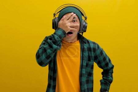 Hombre asiático en un gorro y ropa casual, con auriculares, mira a través de sus dedos en shock después de escuchar algo vergonzoso en su música. Aislado sobre un fondo amarillo.