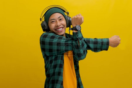 Asian man in a beanie and casual clothes stretches his muscles after work, listening to music on his headphones. A look of focus is on his face. Isolated on a yellow background.