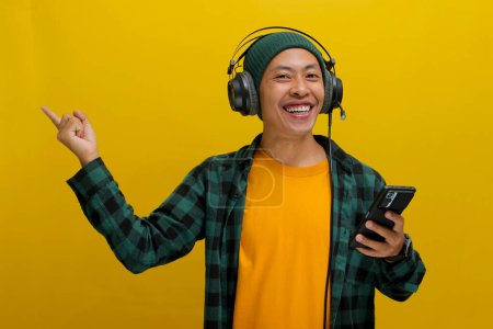 Photo for Asian man in a beanie and casual clothes points his finger towards empty space on the side while listening to music on his headphones. His phone is in his hand. Isolated on a yellow background. - Royalty Free Image
