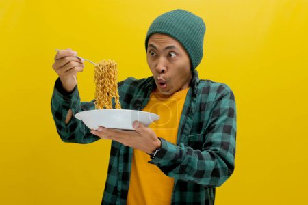 Homme asiatique dans un bonnet et des vêtements décontractés fait une expression surprise et ravie tout en slurping nouilles instantanées avec fourchette. Isolé sur fond jaune.