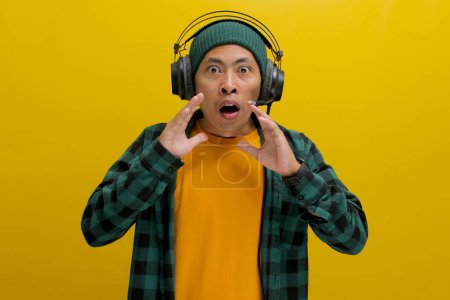 Homme asiatique dans un bonnet et des vêtements décontractés, portant des écouteurs, semble choqué par sa bouche agape après avoir entendu quelque chose de surprenant sur sa musique. Isolé sur fond jaune.