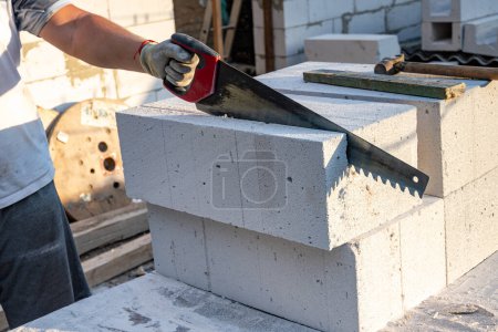 Téléchargez les photos : Un constructeur scie manuellement un bloc de béton aéré avec une scie Construction de briques de béton aéré, blocs. Photo de haute qualité - en image libre de droit