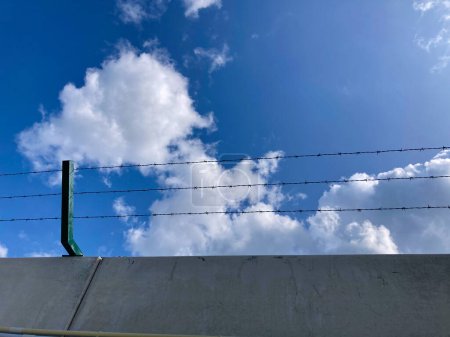 Foto de Muro de hormigón rematado con alambre de púas con cielo azul y nubes blancas por encima - Imagen libre de derechos
