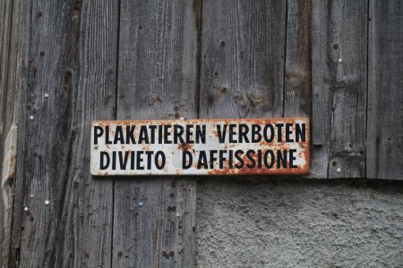 alte verwitterte und verrostete Emailleschilder in deutscher und italienischer Sprache an einer Holzwand. Text lautet "Plakatierung verboten"