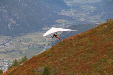 hang glider with white wings and a red and black harness hanging low above the mountain gliding in the air towards the valley below