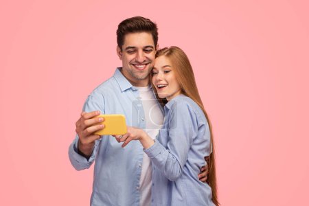 Téléchargez les photos : Joyeux jeune copain et petite amie se câlinant et regardant la vidéo sur téléphone portable sur fond rose - en image libre de droit