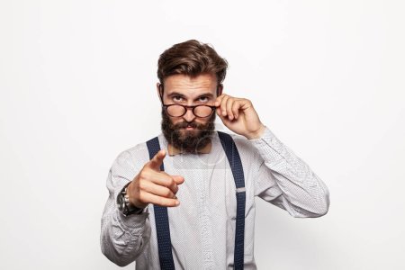 Homme barbu sérieux dans des vêtements élégants ajuster les lunettes et pointant vers la caméra sur fond gris