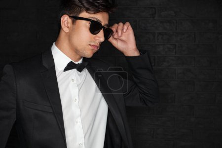 Photo for Young ethnic male in elegant suit adjusting stylish sunglasses and looking away against black brick wall - Royalty Free Image