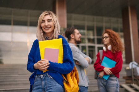 Téléchargez les photos : Femme blonde joyeuse avec sac à dos et papeterie souriant et regardant la caméra tout en se tenant près de parler amis à l'extérieur du bâtiment de l'université en journée - en image libre de droit