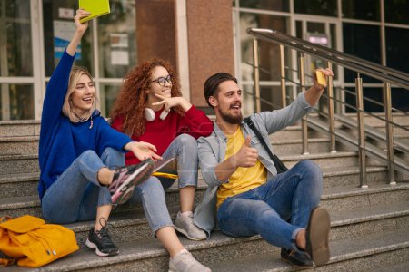 Photo for Delighted young male and female students in casual clothes smiling happily and showing thumb up and two fingers gestures, while taking selfie on smartphone sitting on stairs after successful exams - Royalty Free Image