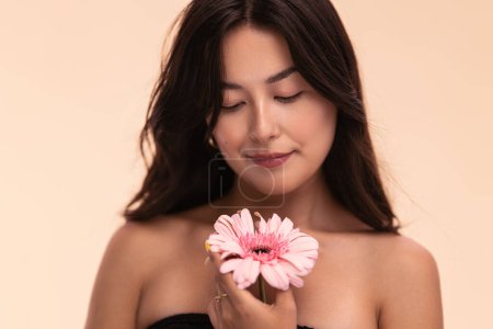 Photo for Asian woman with bare shoulders touching delicate petals of pink gerbera flower against beige background - Royalty Free Image
