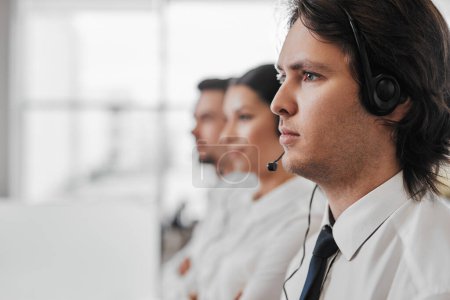Photo for Focused young man with headset looking away near blurred colleagues during work in call center - Royalty Free Image