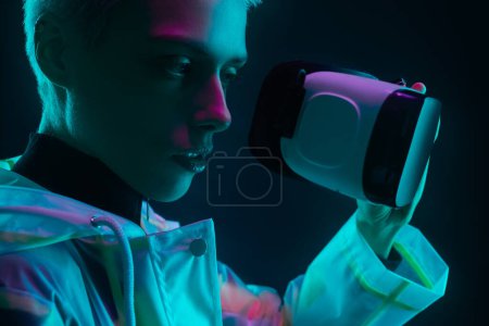 Foto de Mujer andrógina joven en impermeable futurista con casco VR preparándose para explorar el ciberespacio bajo una colorida iluminación de neón sobre fondo oscuro - Imagen libre de derechos