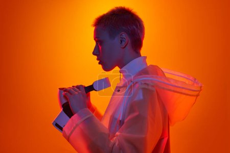 Photo for Side view of of young female gamer in futuristic raincoat with short hair taking off VR goggles, while standing under neon illumination against orange background - Royalty Free Image