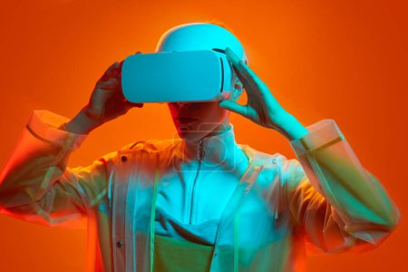 Téléchargez les photos : Jeune femme en tenue futuriste touchant casque VR et explorer le cyberespace sous éclairage au néon coloré sur fond orange - en image libre de droit