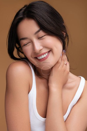 Photo for Cheerful young Asian female smiling with closed eyes and touching neck after hearing flattering compliment against brown background - Royalty Free Image