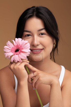 Photo for Glad Asian woman with black hair touching face with fresh gerbera flower and looking at camera with smile against brown background - Royalty Free Image
