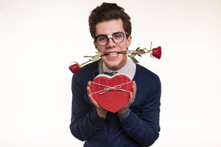 Téléchargez les photos : Jeune homme dans des lunettes tenant des roses dans les dents et montrant boîte cadeau en forme de coeur pendant la date romantique sur fond blanc - en image libre de droit