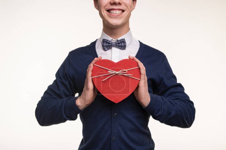 Téléchargez les photos : Crop joyeux jeune homme romantique en chemise blanche et noeud papillon souriant joyeusement tout en démontrant boîte cadeau en forme de coeur rouge sur fond blanc - en image libre de droit