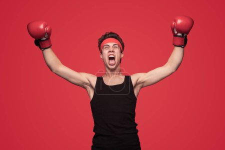 Foto de Joven deportista con camiseta sin mangas y guantes levantando brazos y gritando mientras celebra la victoria en el combate de boxeo contra fondo rojo - Imagen libre de derechos
