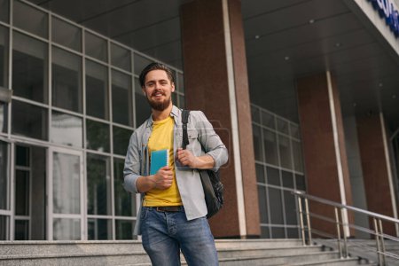 Téléchargez les photos : Angle bas de jeune étudiant masculin barbu positif en vêtements décontractés avec sac à dos et cahier, souriant et regardant la caméra tout en se tenant debout sur les escaliers du bâtiment de l'université - en image libre de droit