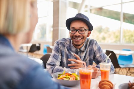 Téléchargez les photos : Heureux homme afro-américain en chemise à carreaux avec des lunettes et un chapeau souriant et parlant avec sa petite amie pendant le déjeuner au restaurant - en image libre de droit