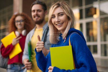 Téléchargez les photos : Jeune femme blonde positive en sweat à capuche bleu avec carnet vert regardant la caméra avec le sourire et le pouce levé, tout en se tenant près des amis après l'inscription sur le campus universitaire - en image libre de droit