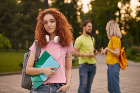 Photo for Young female millennial with curly red hair in casual clothes and headphone son neck, standing in park with folder and notebook near groupmates talking after - Royalty Free Image