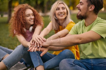 Foto de Positivo jóvenes amigos masculinos y femeninos en ropa casual sonriendo y apilando las manos mientras se sientan juntos en el césped cubierto de hierba en el parque - Imagen libre de derechos