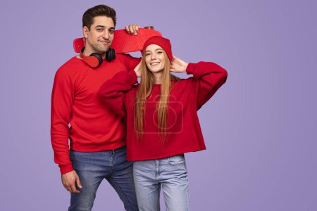 Photo for Happy young woman in stylish clothes touching hat and looking at camera with smile while standing near boyfriend with penny board against violet background - Royalty Free Image